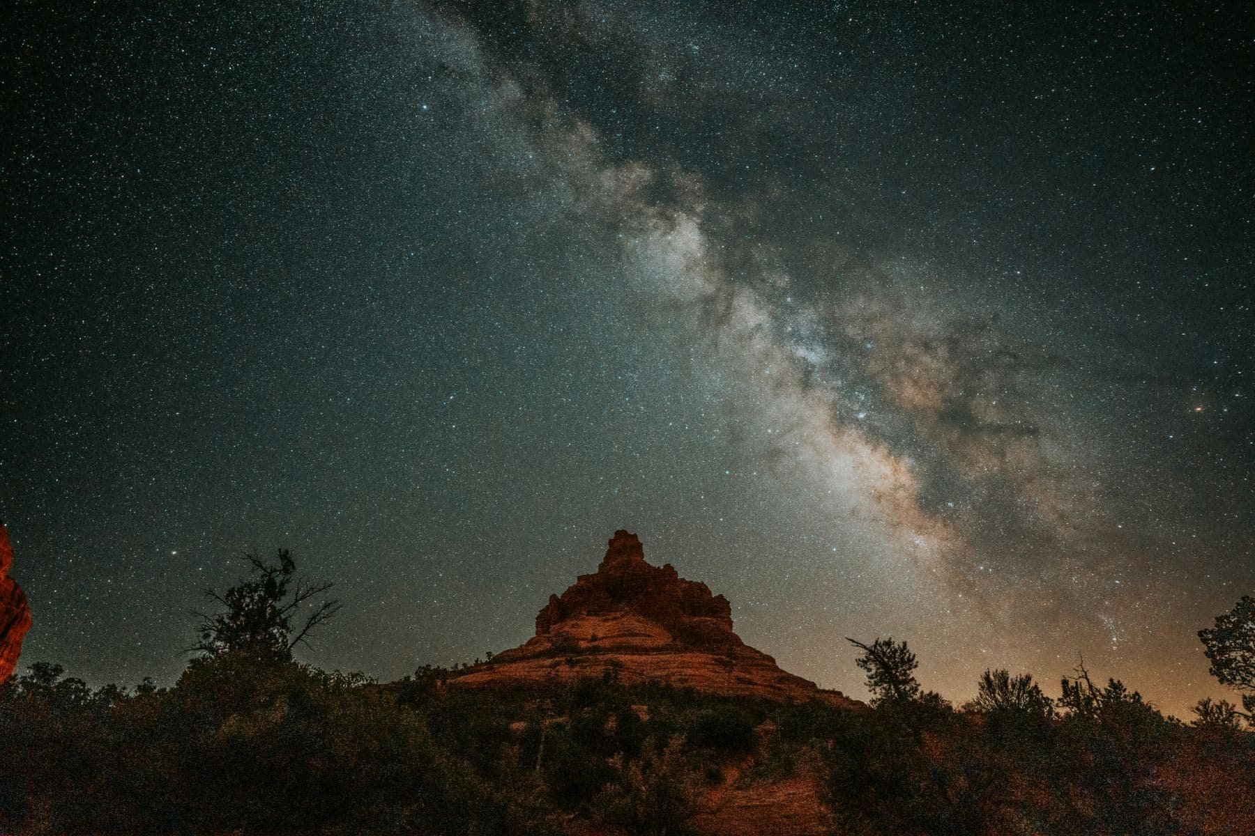 The Milky Way arching over a red sandstone butte and dark desert silhouette where prickly pear cactus grows